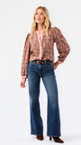 A smiling blonde woman models a long-sleeve patterned blouse with pink trim and blue jeans against a plain white studio background.