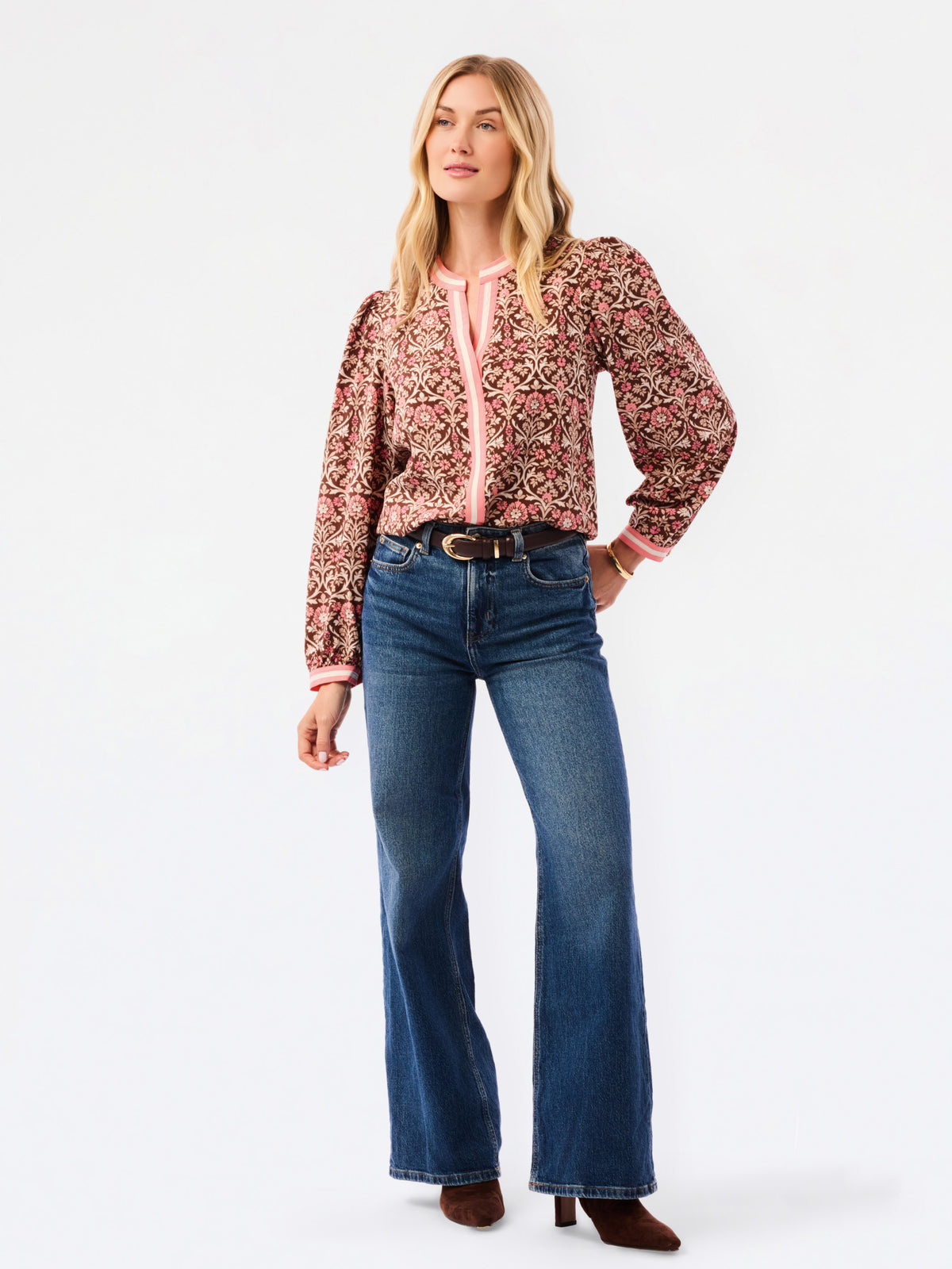 A smiling blonde woman models a long-sleeve patterned blouse with pink trim and blue jeans against a plain white studio background.