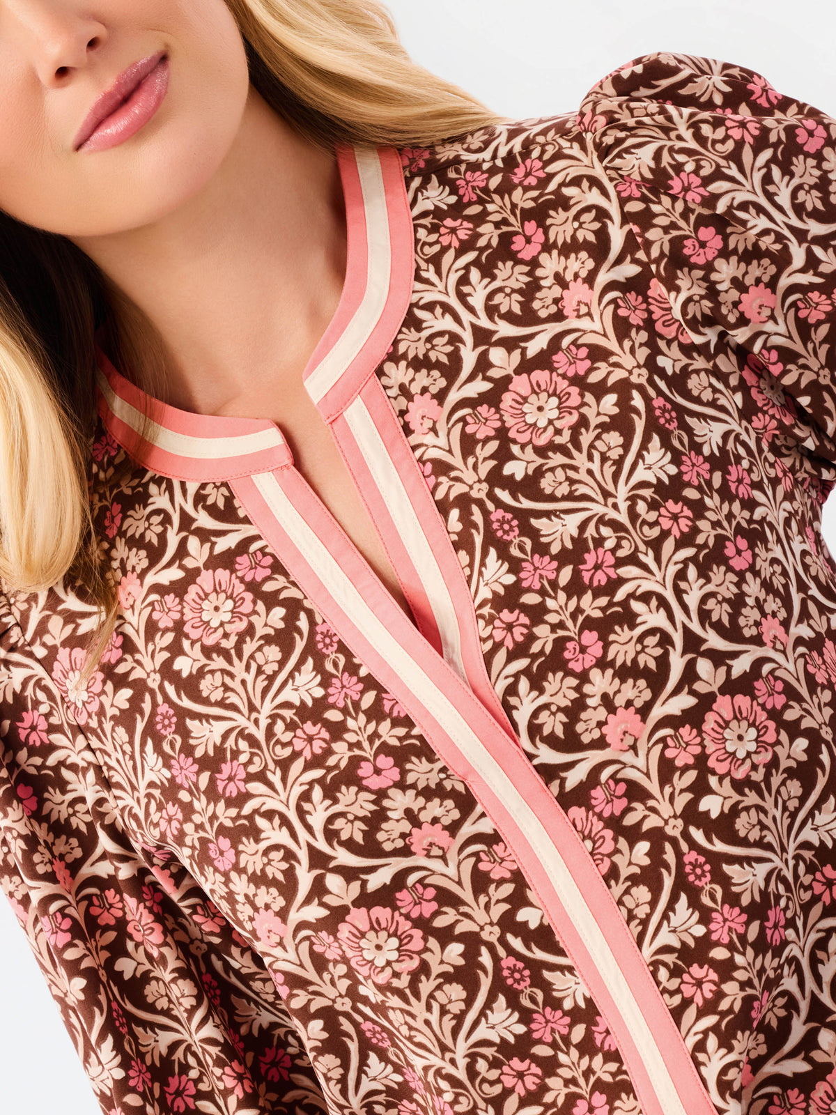 A smiling blonde woman models a long-sleeve patterned blouse with pink trim and blue jeans against a plain white studio background.