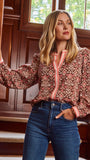 A smiling blonde woman models a long-sleeve patterned blouse with pink trim and blue jeans 