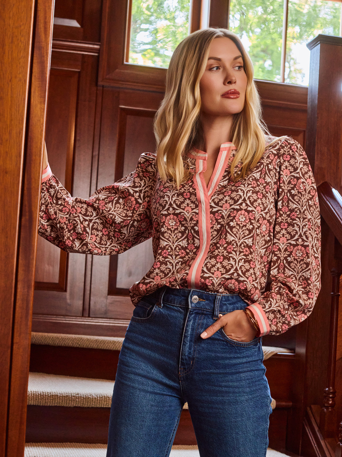 A smiling blonde woman models a long-sleeve patterned blouse with pink trim and blue jeans 