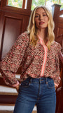 A smiling blonde woman models a long-sleeve patterned blouse with pink trim and blue jeans 