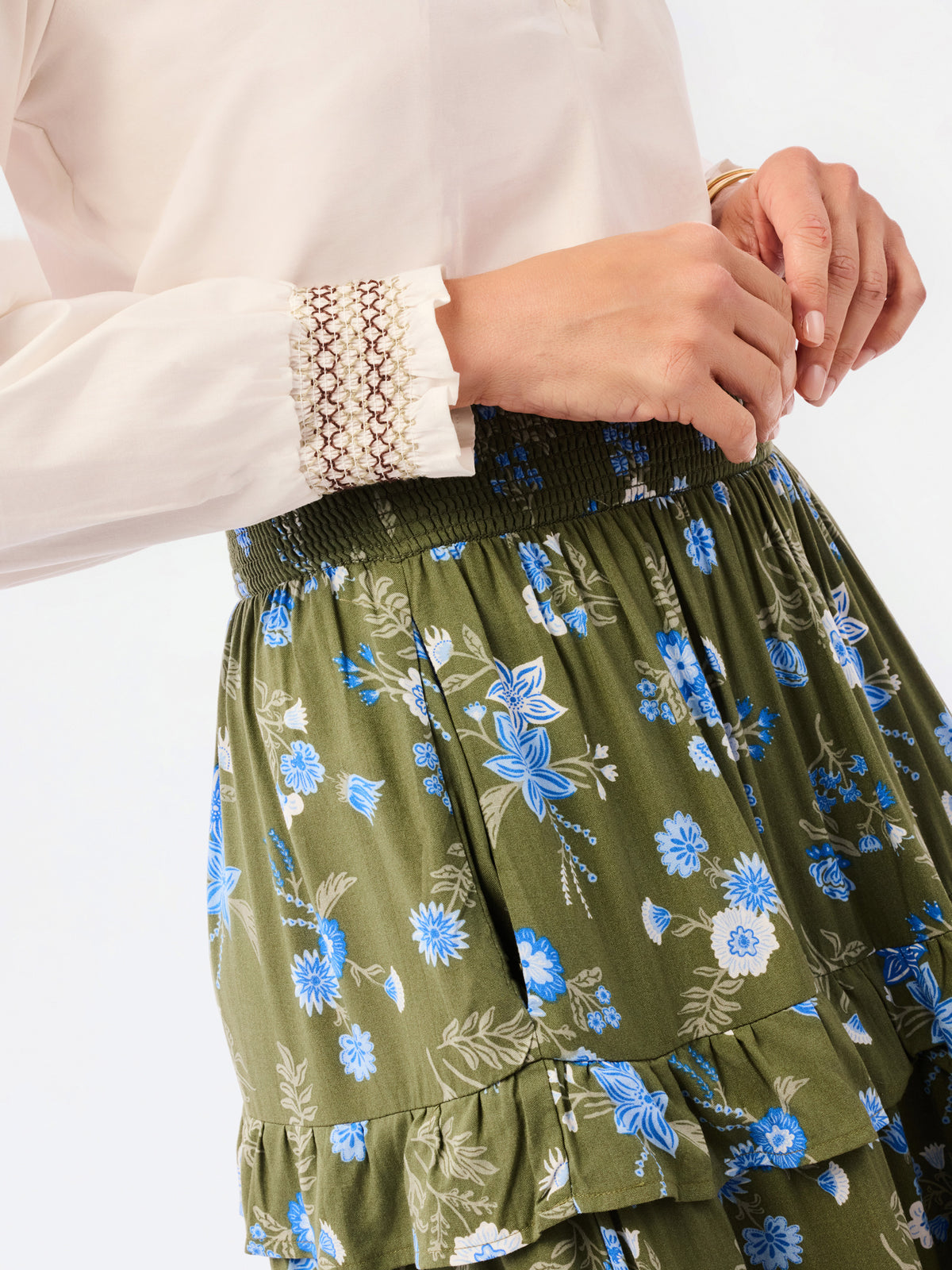 Close-up of a person wearing an olive green tiered skirt with a blue and white floral print, paired with a cream blouse featuring a lace cuff.