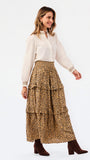 A woman modeling a high-waisted, tiered leopard-print maxi skirt paired with a cream blouse and brown ankle boots, set against a plain white studio background.