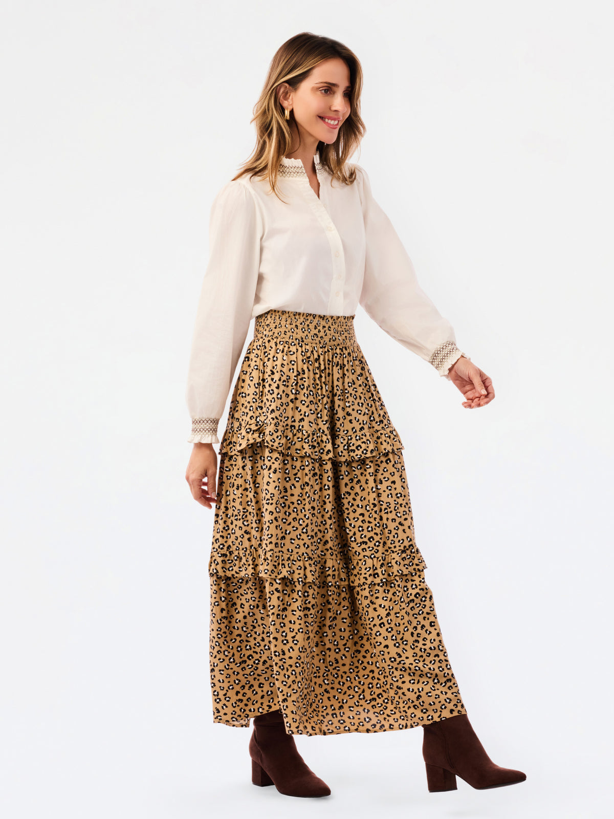 A woman modeling a high-waisted, tiered leopard-print maxi skirt paired with a cream blouse and brown ankle boots, set against a plain white studio background.