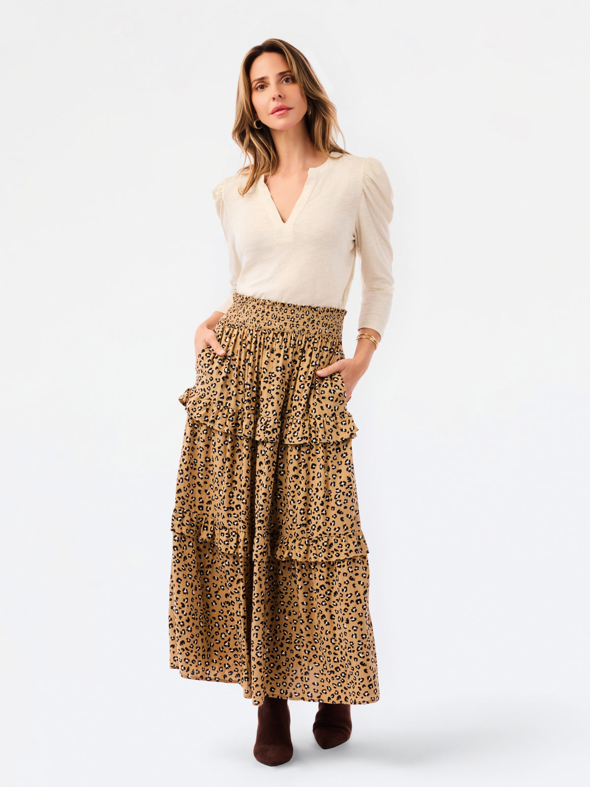 A woman modeling a long tiered skirt with a brown and black leopard print, paired with a cream-colored long-sleeve top and dark brown boots, against a plain white studio background.
