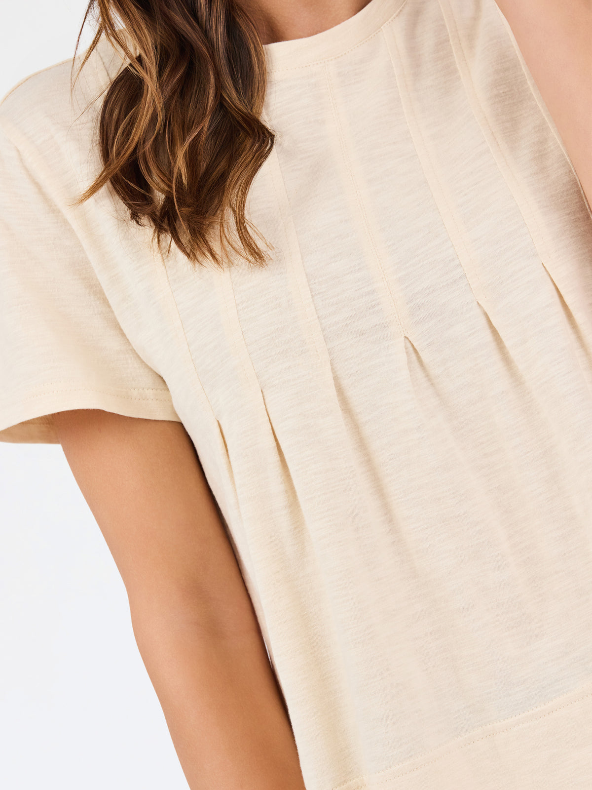 Close-up of a person wearing a short-sleeve cream-colored top with vertical pleated detailing on a light background.