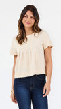 A woman modeling a short-sleeve cream-colored knit top with a gathered, pleated front, paired with dark blue jeans, against a plain white studio background.