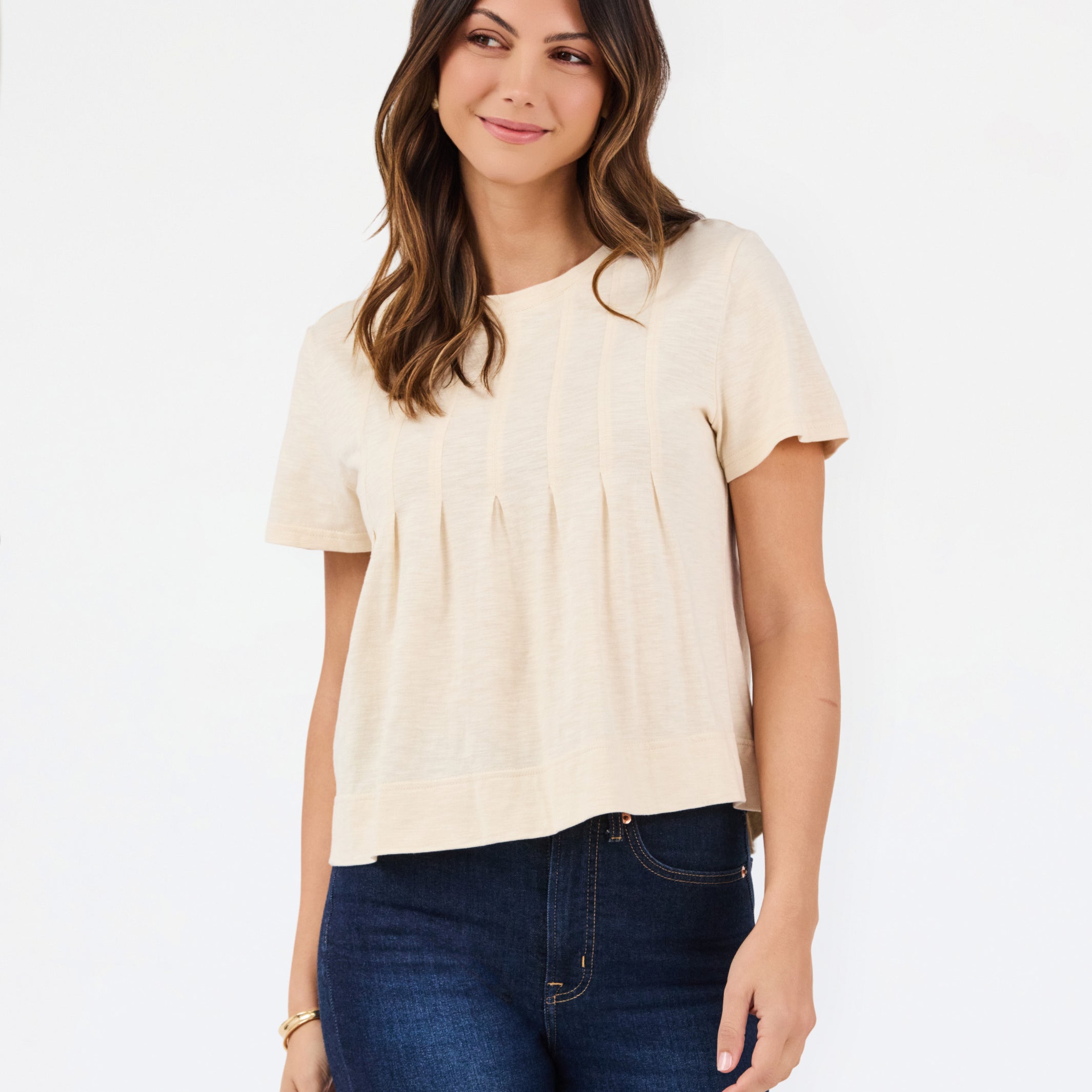 A woman modeling a short-sleeve cream-colored knit top with a gathered, pleated front, paired with dark blue jeans, against a plain white studio background.