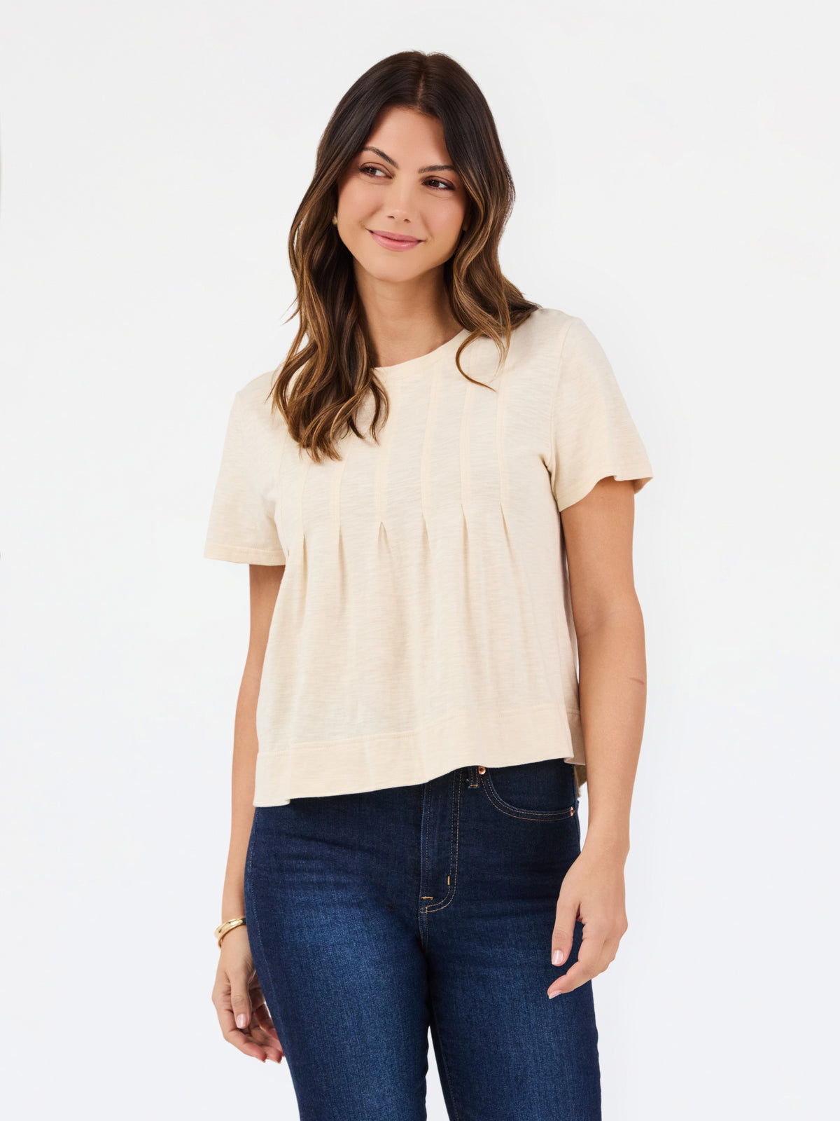 A woman modeling a short-sleeve cream-colored knit top with a gathered, pleated front, paired with dark blue jeans, against a plain white studio background.