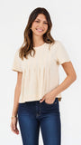 A woman modeling a short-sleeve cream-colored knit top with pleated detailing on the front, paired with dark blue jeans, against a plain white studio background.
