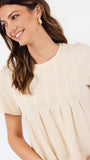 A woman wearing a short-sleeve cream-colored top with vertical pleated detailing on the chest and a gathered waist, photographed against a clean white background.