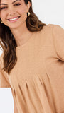 A woman modeling a short-sleeve, crew-neck top in a light tan or beige color, paired with dark blue jeans, against a plain white studio background.