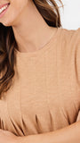 A woman modeling a short-sleeve, crew-neck top in a light tan or beige color, paired with dark blue jeans, against a plain white studio background.