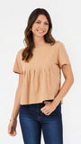 A woman modeling a short-sleeve, crew-neck top in a light tan or beige color, paired with dark blue jeans, against a plain white studio background.