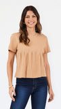 A woman modeling a short-sleeve, crew-neck top in a light tan or beige color, paired with dark blue jeans, against a plain white studio background.