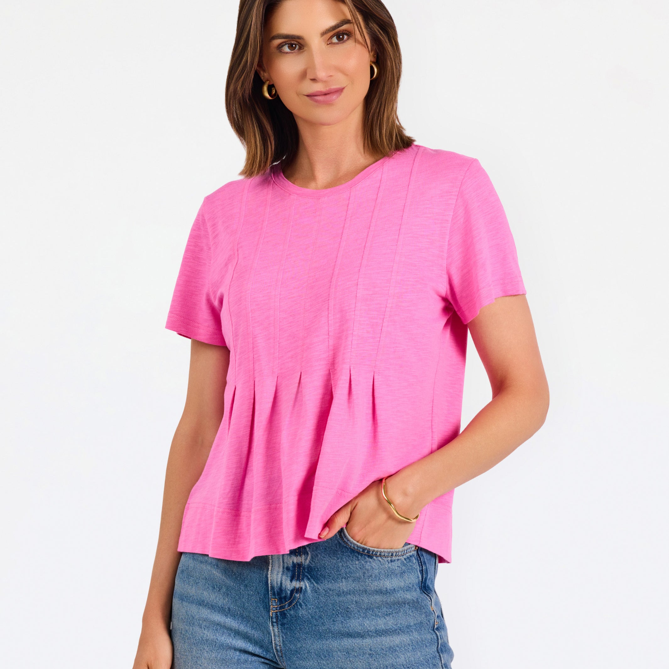 A woman modeling a bright pink short-sleeve top with a gathered peplum hem, paired with blue jeans, against a plain white studio background.