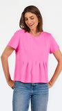 A woman modeling a bright pink short-sleeve top with a gathered peplum hem, paired with blue jeans, against a plain white studio background.