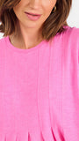 A woman modeling a bright pink short-sleeve top with a gathered peplum hem, paired with blue jeans, against a plain white studio background.