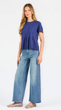 A woman modeling a short-sleeve peplum top in a heathered periwinkle blue color, paired with light-wash blue jeans, against a plain white studio background.