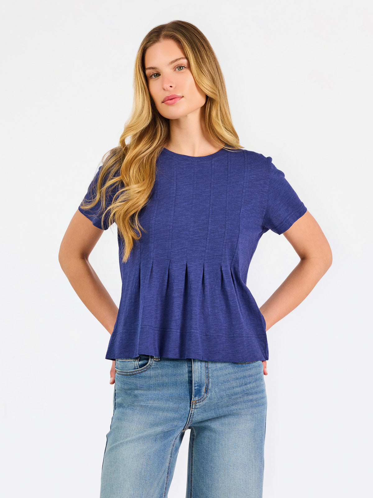 A woman modeling a short-sleeve peplum top in a heathered periwinkle blue color, paired with light-wash blue jeans, against a plain white studio background.