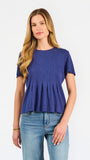 A woman modeling a short-sleeve peplum top in a heathered periwinkle blue color, paired with light-wash blue jeans, against a plain white studio background.