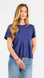 A woman modeling a short-sleeve peplum top in a heathered periwinkle blue color, paired with light-wash blue jeans, against a plain white studio background.