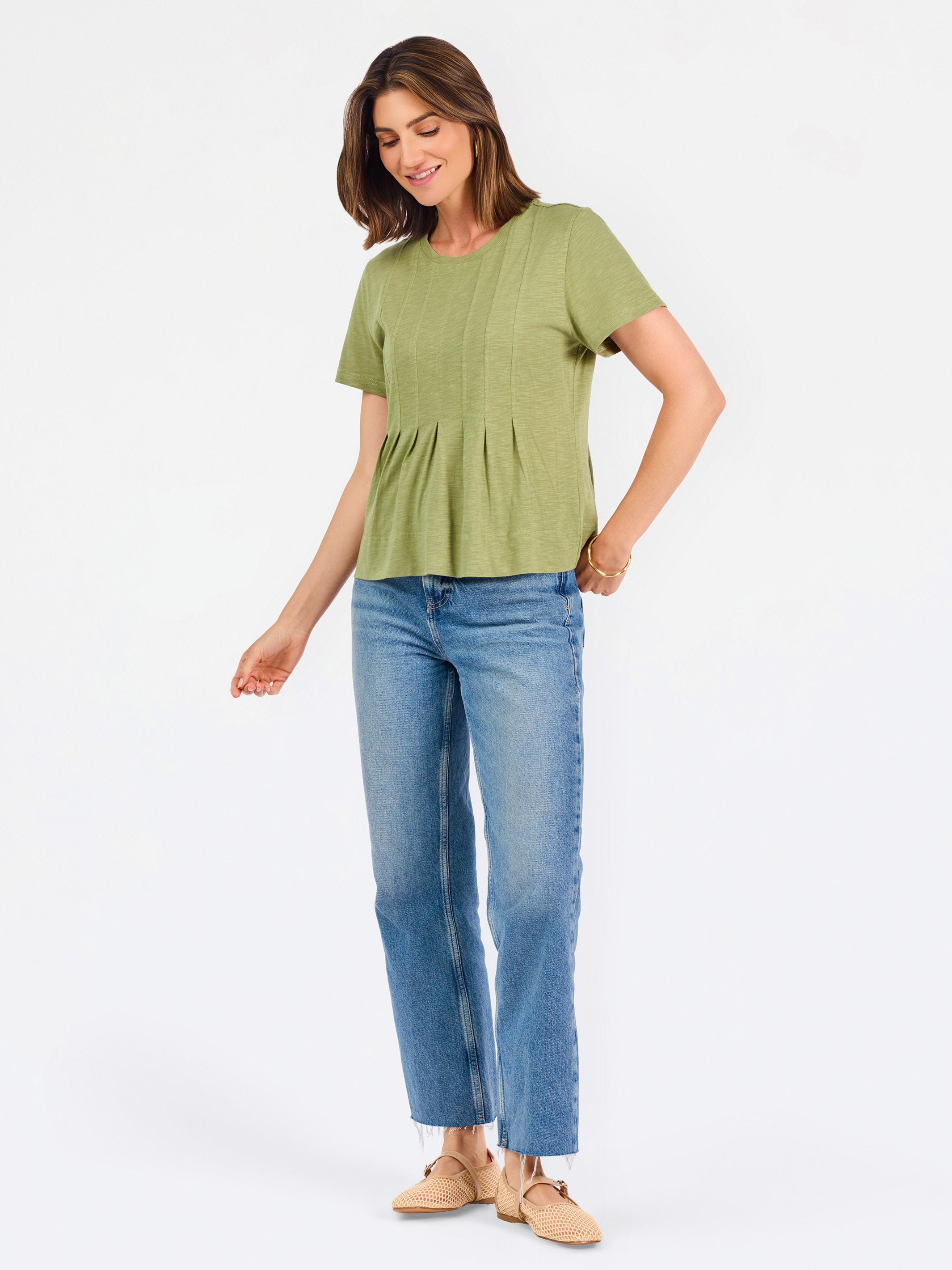 A woman wearing a sage green short-sleeve top with a gathered peplum hem, paired with blue jeans, against a plain white background.