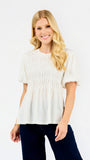 A smiling blonde woman wearing a short-sleeve white peplum top with smocked detailing and dark navy pants, standing against a plain white studio background.