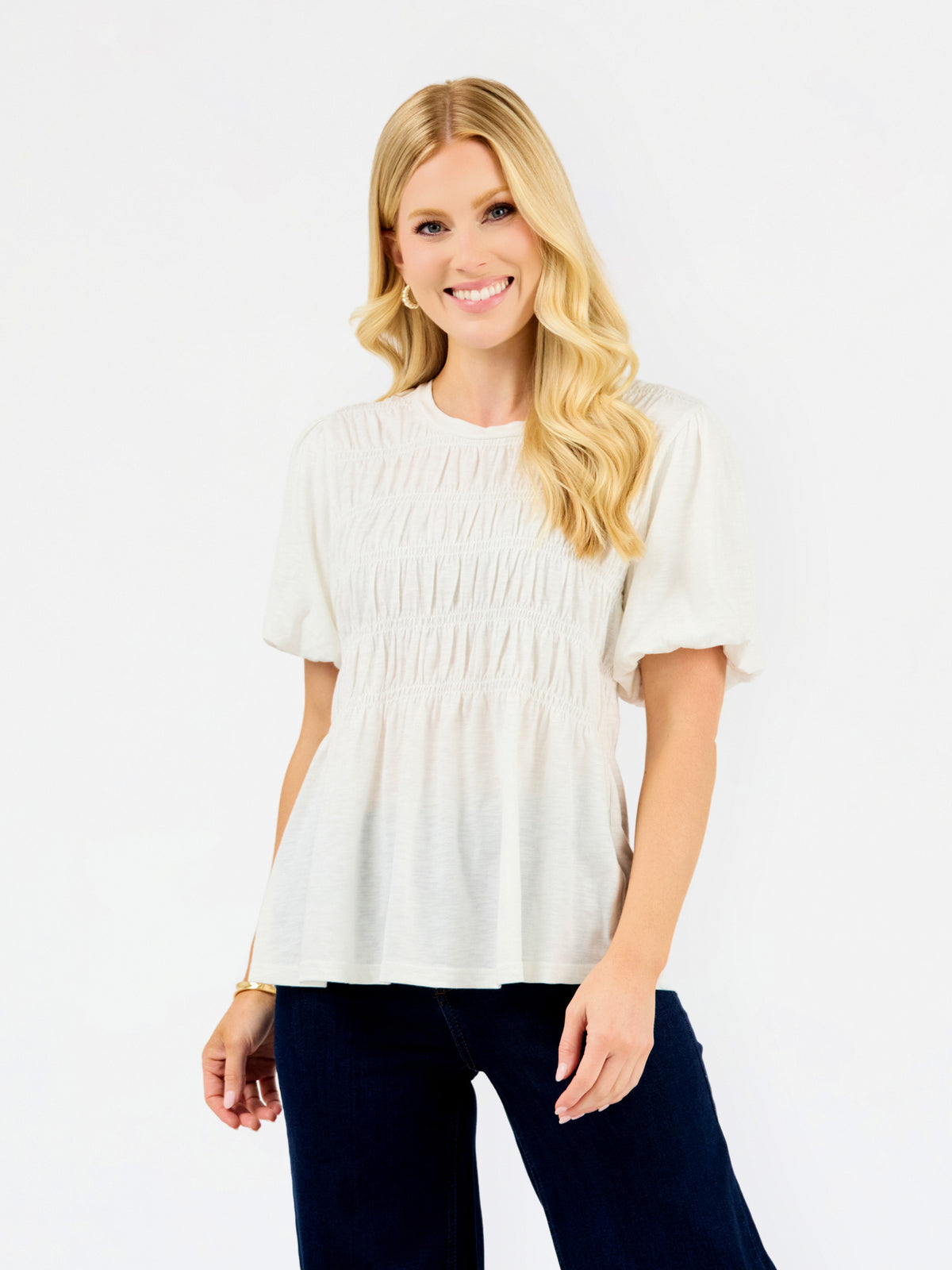A smiling blonde woman wearing a short-sleeve white peplum top with smocked detailing and dark navy pants, standing against a plain white studio background.