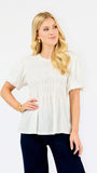 A blonde woman wearing a short-sleeve white smocked blouse and dark navy pants, posing against a solid white studio background.