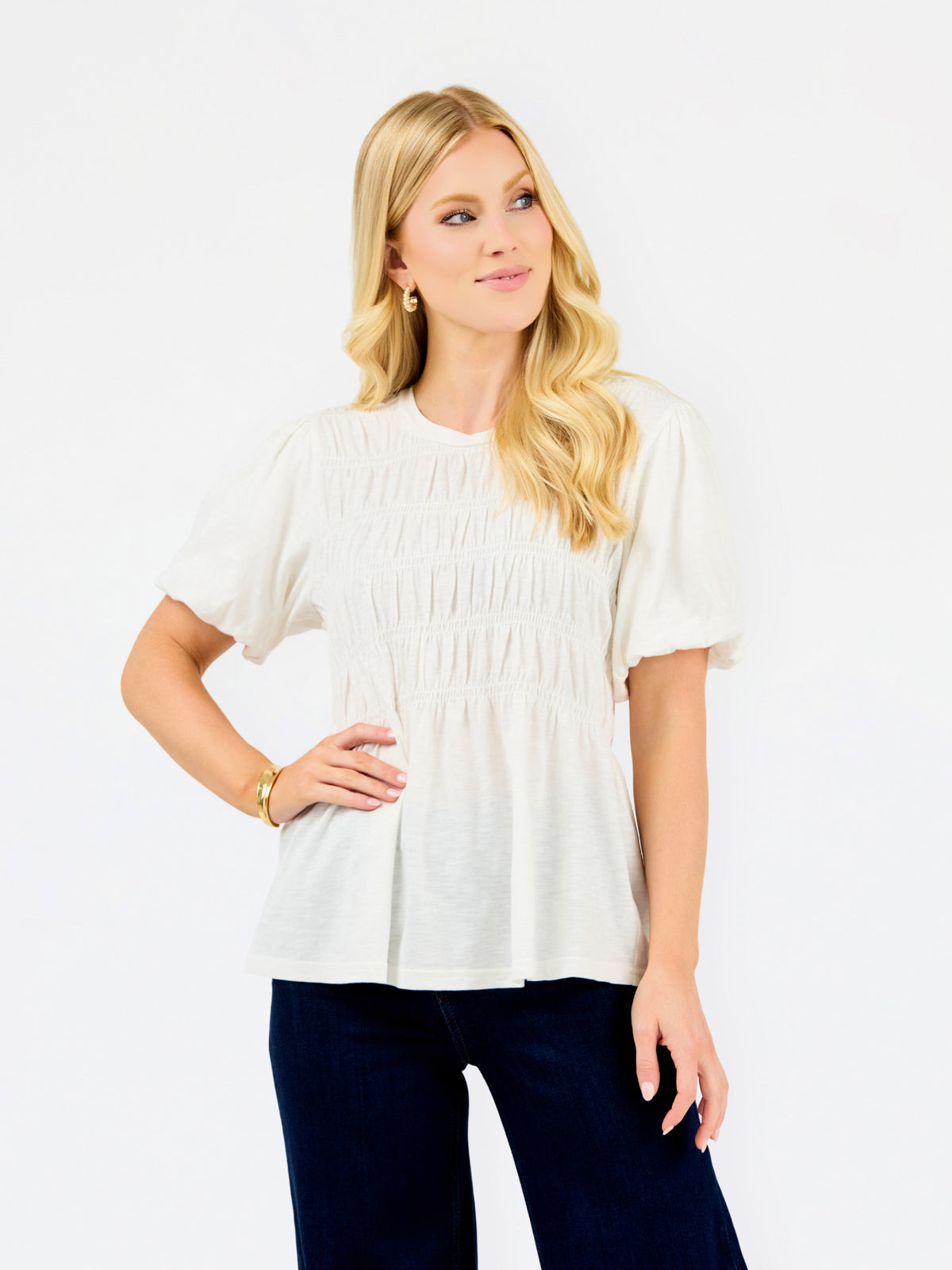 A blonde woman wearing a short-sleeve white smocked blouse and dark navy pants, posing against a solid white studio background.