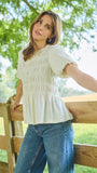 A smiling brunette woman wearing a short-sleeve white peplum top with smocked detailing and jean leaning againzst a fence. 
