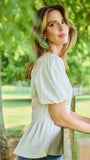 A woman wearing a light sage green puff-sleeve blouse and blue jeans, leaning against a wooden fence in a sunny, green park setting.