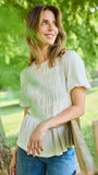 A woman wearing a cream-colored, short-sleeved smocked blouse and blue jeans, standing outdoors in front of a lush green tree.