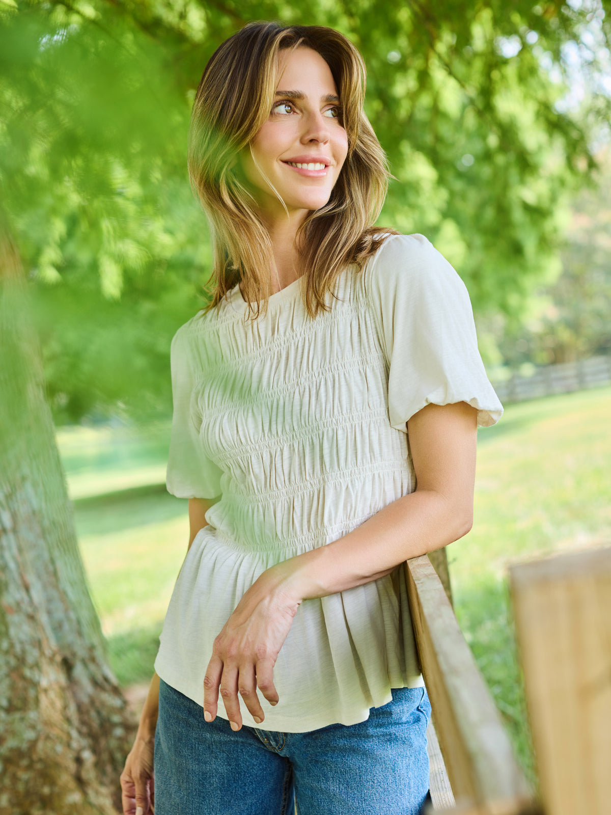 A woman wearing a cream-colored, short-sleeved smocked blouse and blue jeans, standing outdoors in front of a lush green tree.