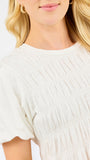 Close-up of a woman wearing a cream-colored short-sleeve top with a smocked bodice and gathered sleeves against a plain white background.