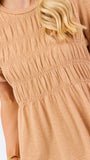 A woman with blonde hair wearing a tan short-sleeve peplum top with smocked detailing and blue jeans, standing against a plain white background.
