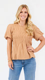 A woman with blonde hair wearing a tan short-sleeve peplum top with smocked detailing and blue jeans, standing against a plain white background.