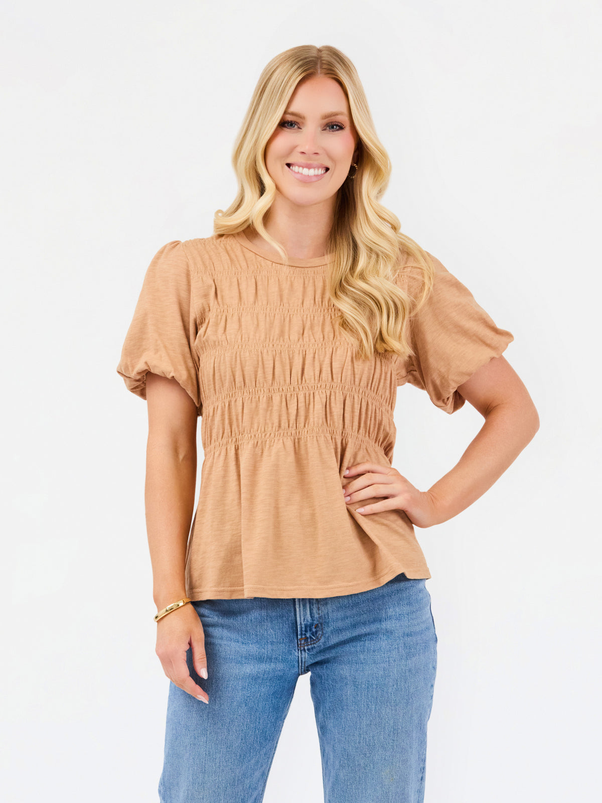 A woman with blonde hair wearing a tan short-sleeve peplum top with smocked detailing and blue jeans, standing against a plain white background.