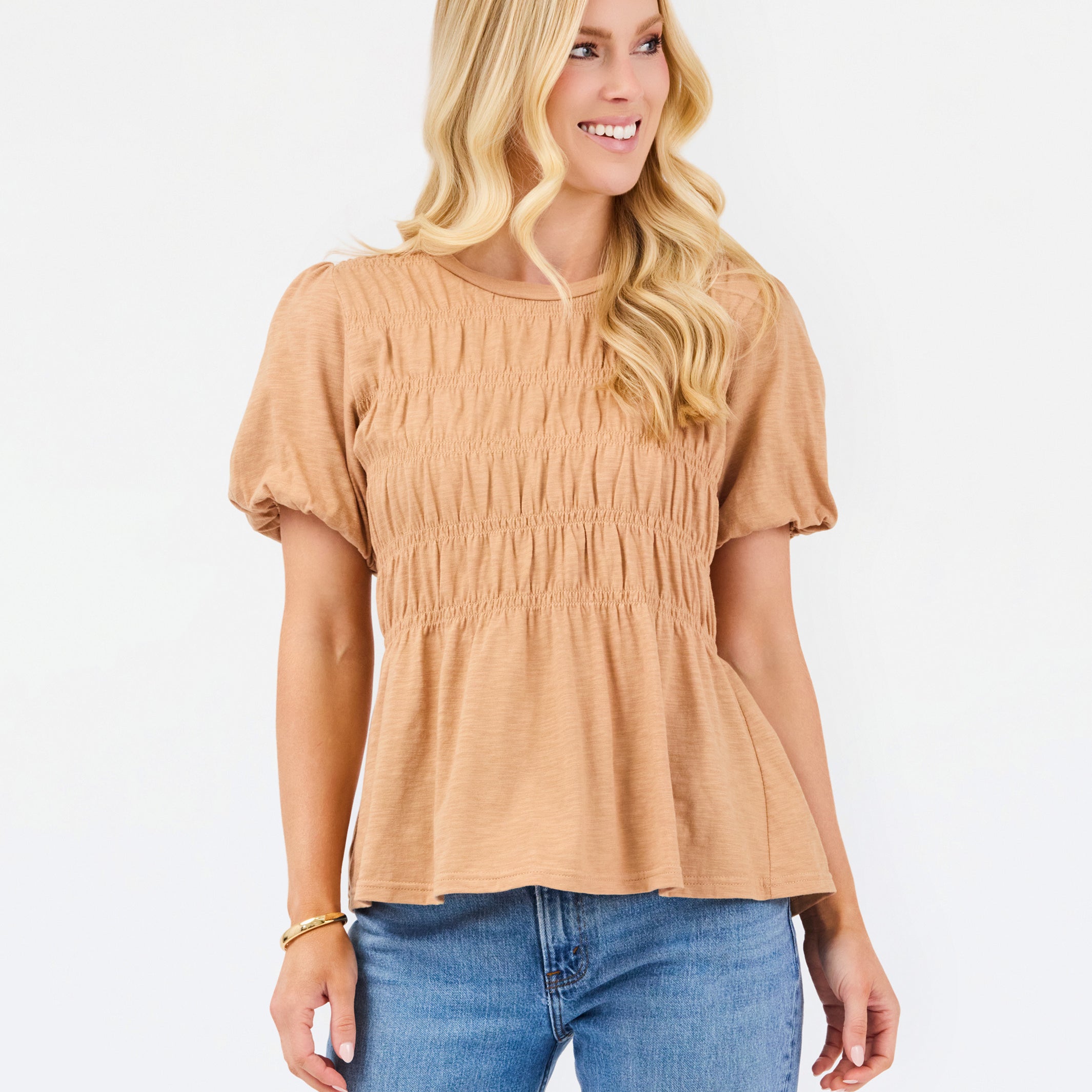 A woman with blonde hair wearing a tan short-sleeve peplum top with smocked detailing and blue jeans, standing against a plain white background.