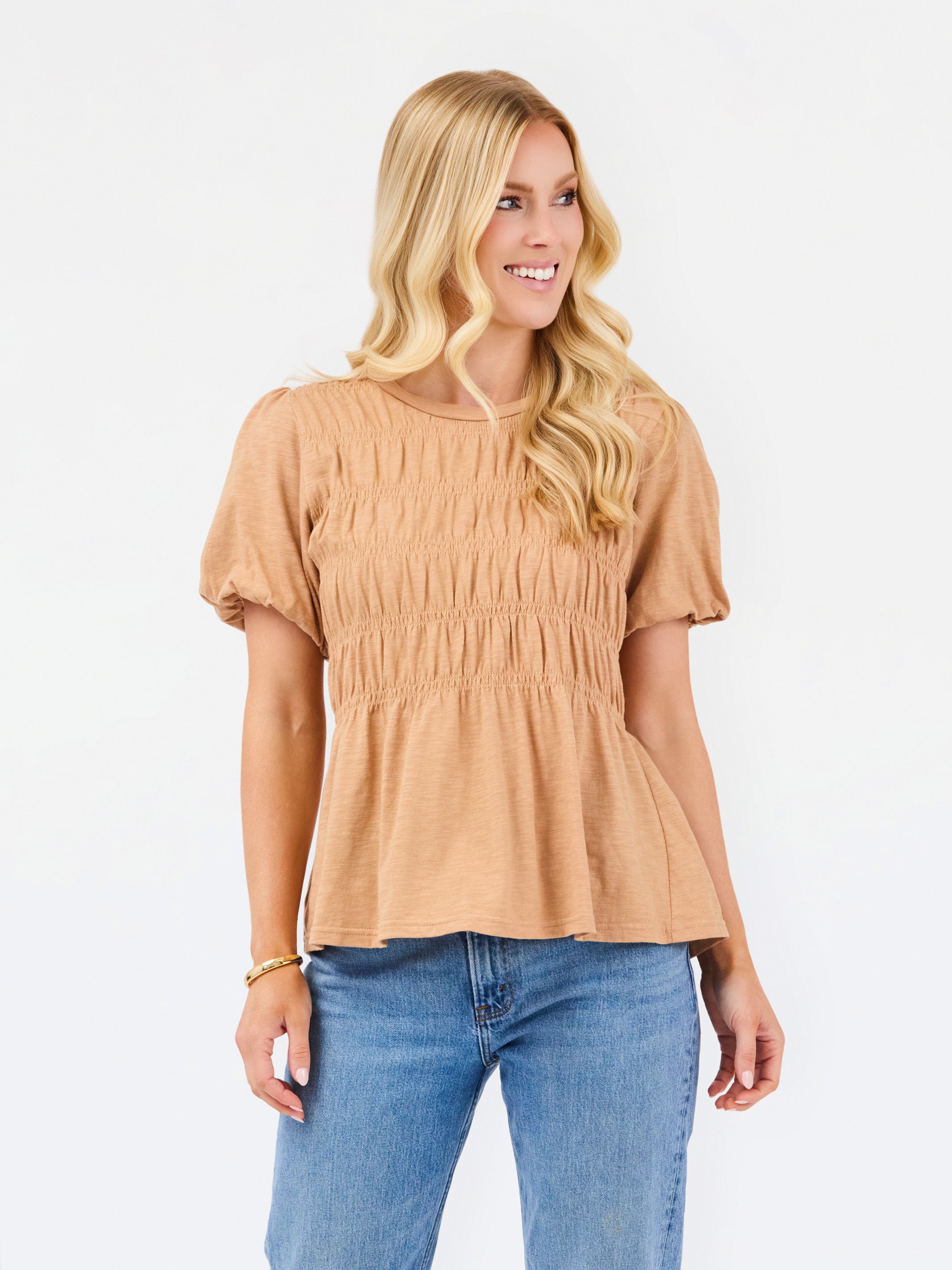 A woman with blonde hair wearing a tan short-sleeve peplum top with smocked detailing and blue jeans, standing against a plain white background.