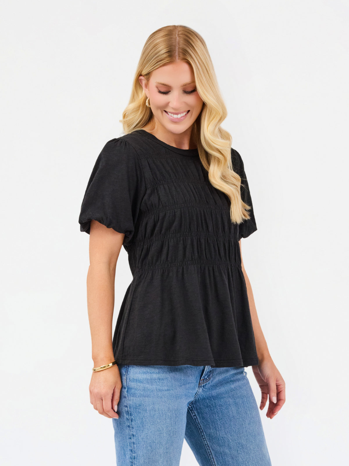 A woman with blonde hair wearing a black short-sleeve peplum top and blue jeans, standing against a plain white background.