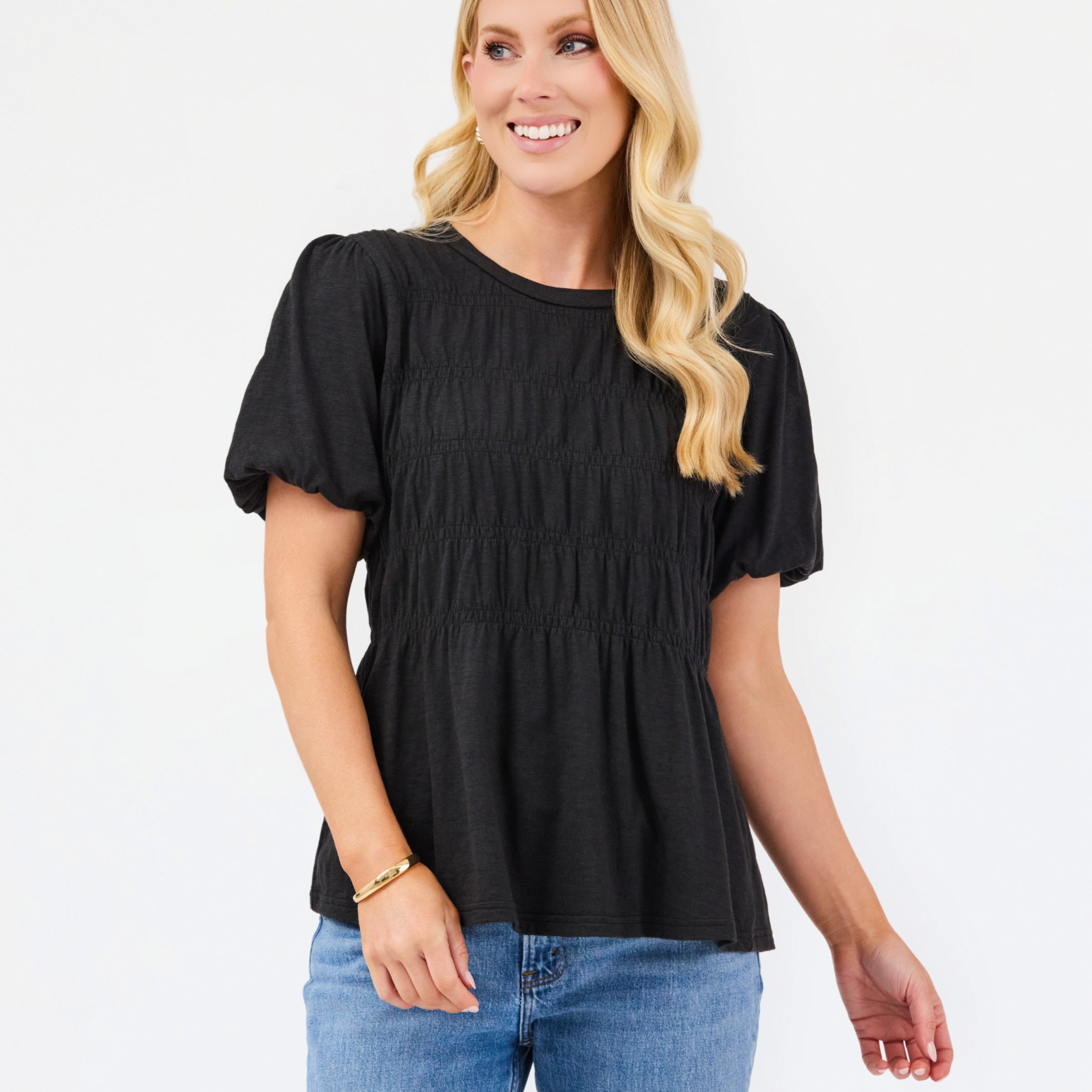 A woman modeling a black short-sleeve peplum top with puff sleeves and a smocked bodice, paired with blue jeans, against a plain white studio background.