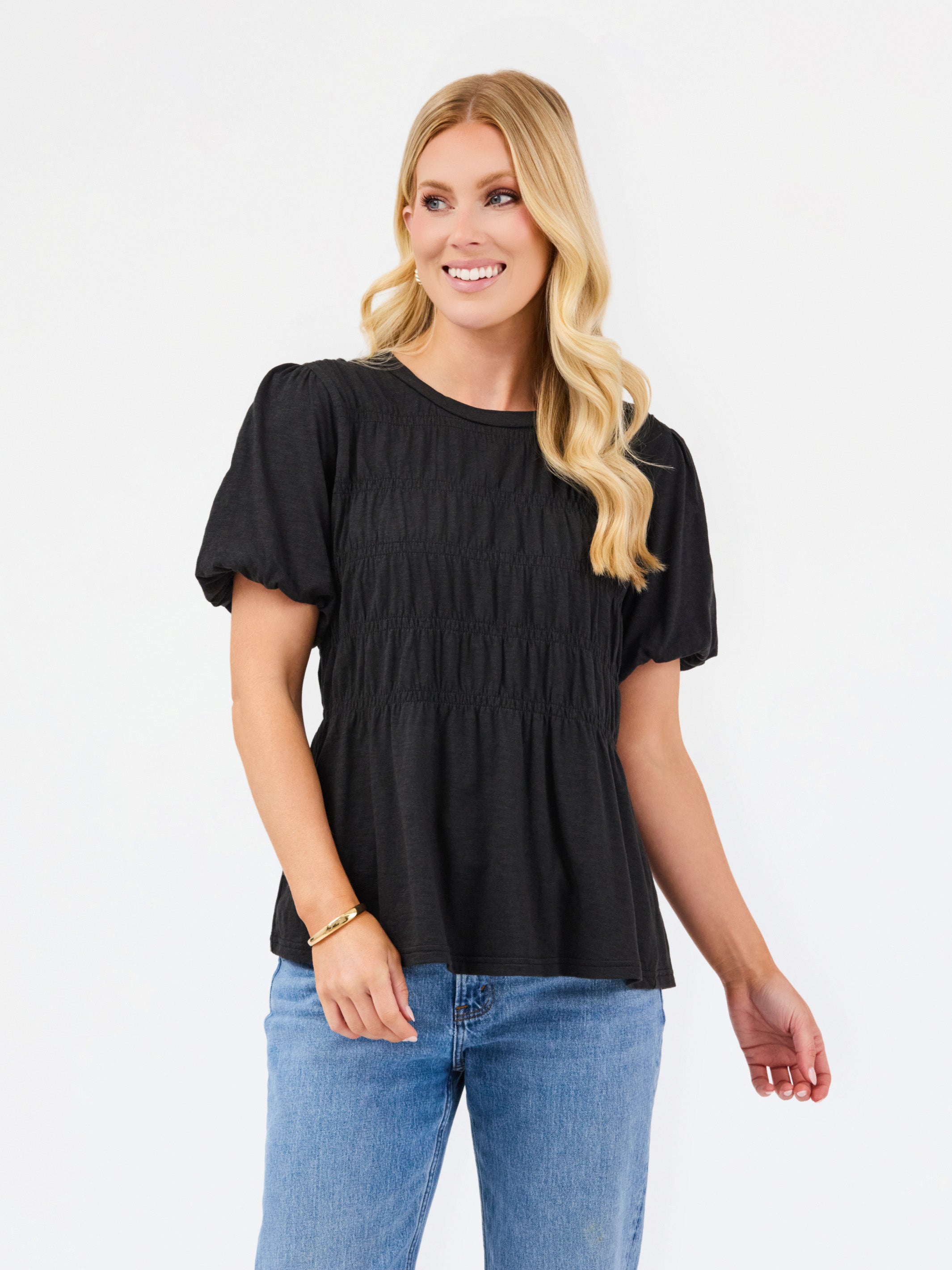 A woman modeling a black short-sleeve peplum top with puff sleeves and a smocked bodice, paired with blue jeans, against a plain white studio background.