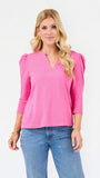A blonde woman wearing a bright pink three-quarter sleeve top with puffed shoulders and blue jeans, standing against a plain white studio background.