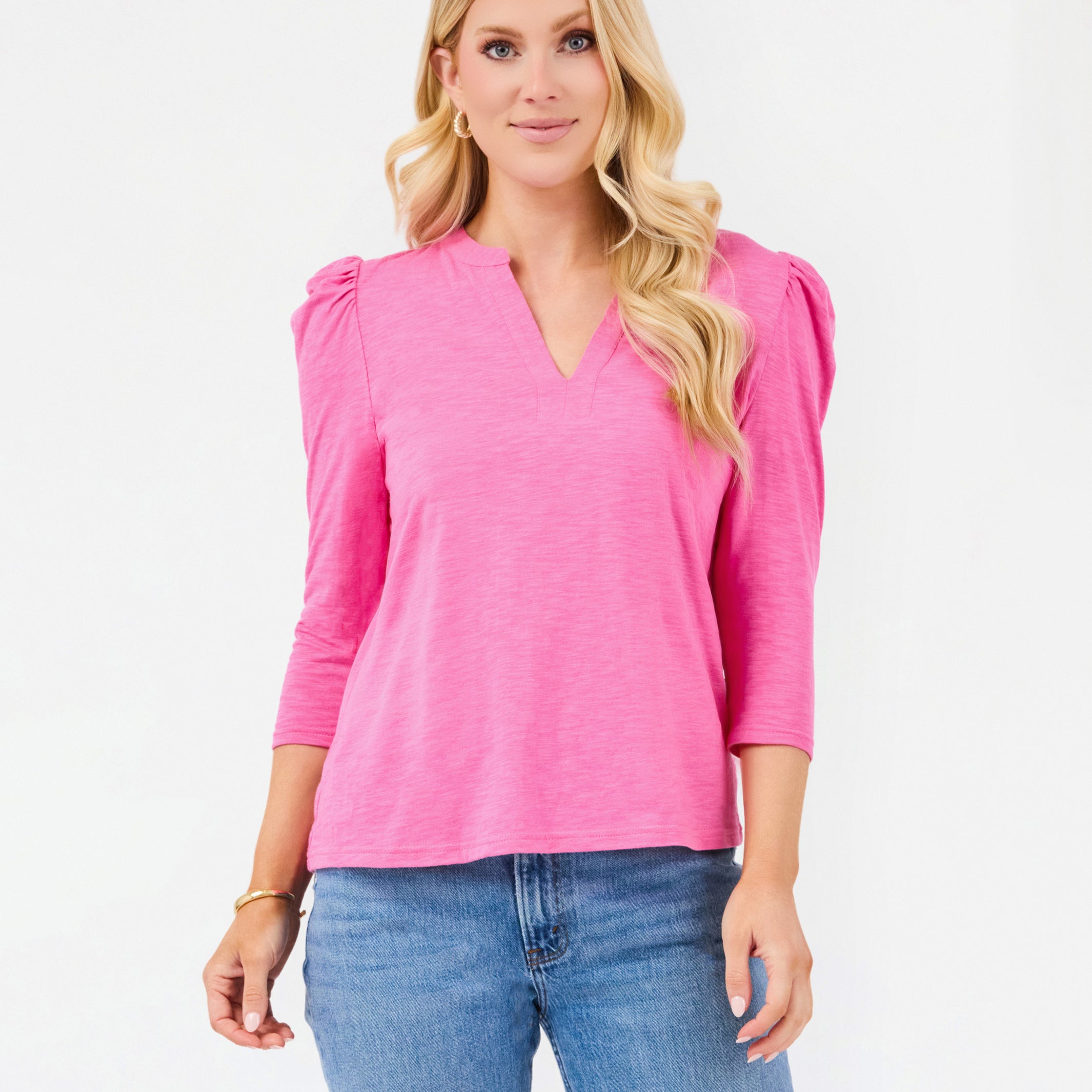 A blonde woman wearing a bright pink three-quarter sleeve top with puffed shoulders and blue jeans, standing against a plain white studio background.