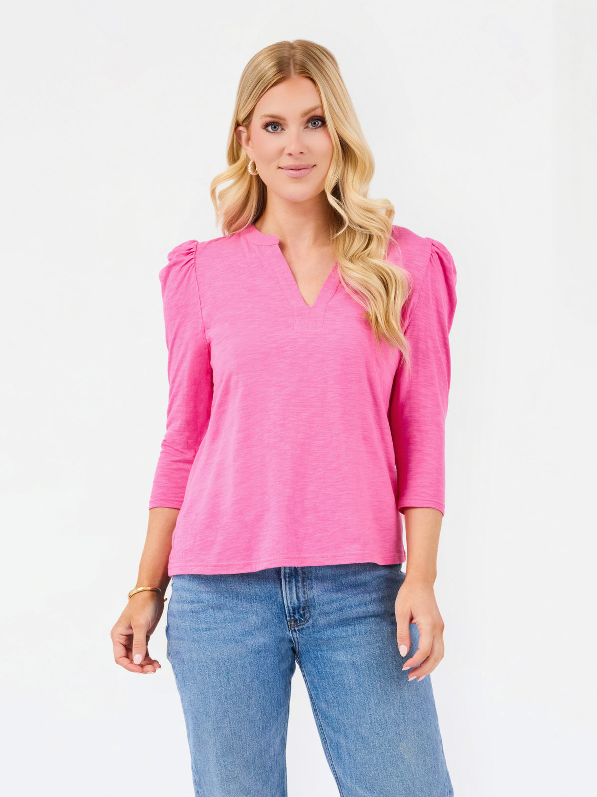 A blonde woman wearing a bright pink three-quarter sleeve top with puffed shoulders and blue jeans, standing against a plain white studio background.