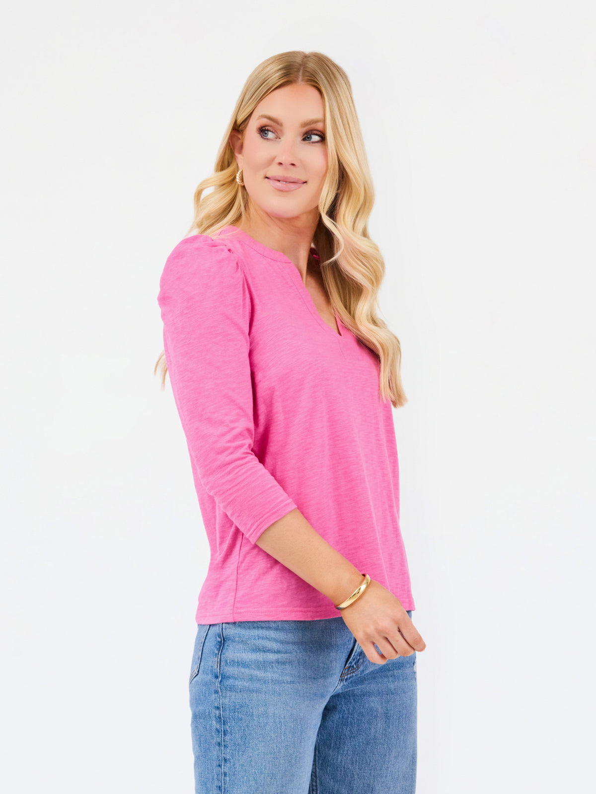 A woman modeling a bright pink three-quarter sleeve top with a split neckline, paired with blue jeans, against a plain white studio background.