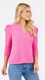 A blonde woman wearing a bright pink three-quarter sleeve top with puffed shoulders and a V-neckline, paired with blue jeans, photographed against a plain white studio background.